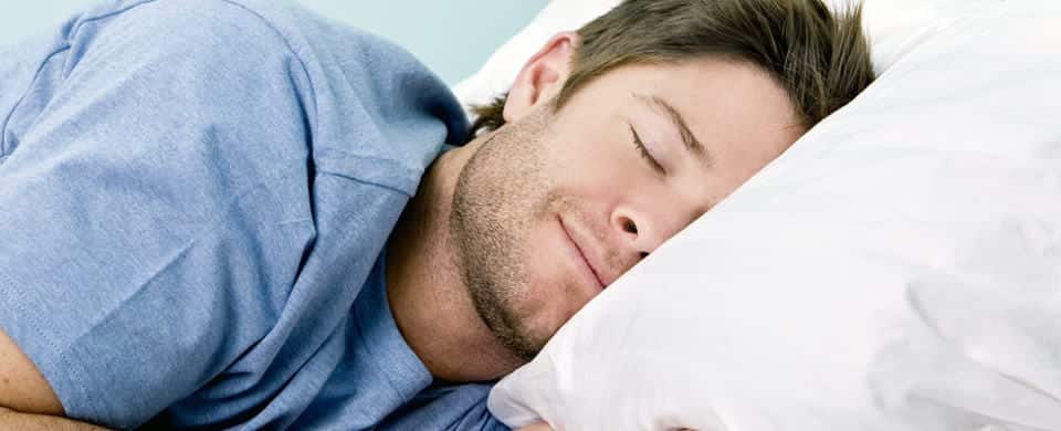 A man with short hair and a light stubble is smiling while sleeping on a white pillow, wearing a light blue t-shirt.