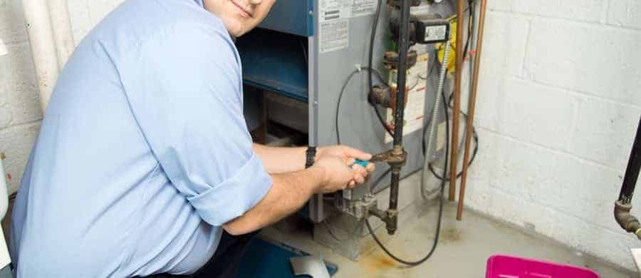 A person in a blue shirt kneels to adjust a valve on a furnace in a basement. Tools and a pink container are scattered nearby.