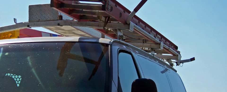 A white van with a red ladder secured on its roof rack, under a clear blue sky.
