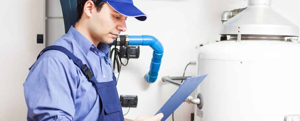 A technician in blue uniform examines a water heater, holding a clipboard.