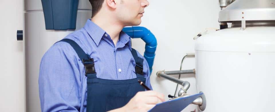 A worker in blue overalls inspects a water heater, holding a clipboard and pen.