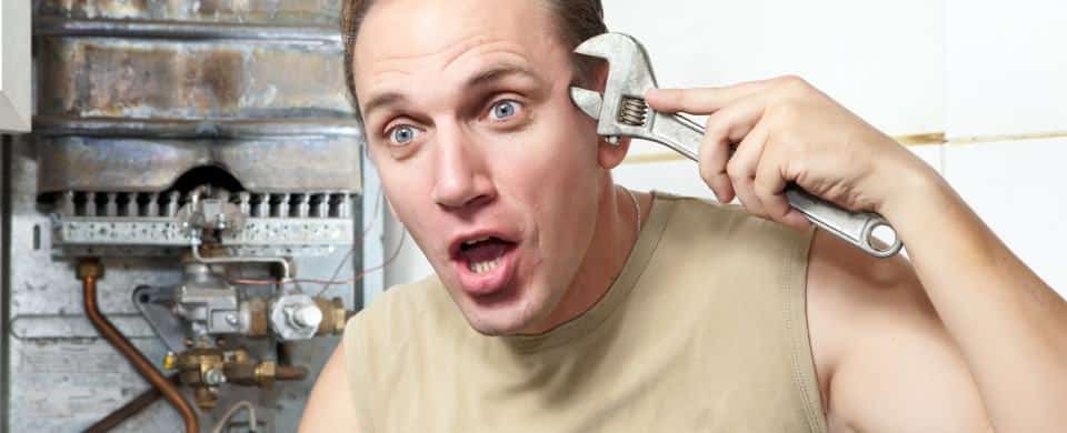 A man holds a wrench to his head, making a surprised or shocked face, with a boiler and pipes in the background—perfectly capturing the frustration of unexpected water heater problems.