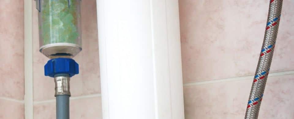 Close-up of plumbing pipes and a water filter with a blue valve against a tiled wall.