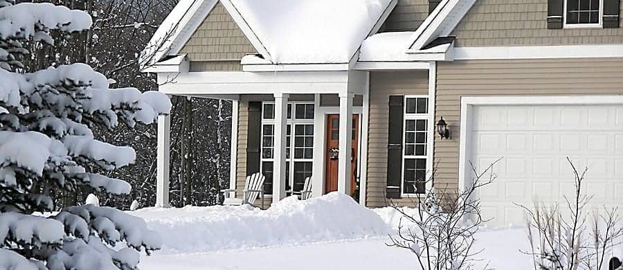 Snow-covered house with a front porch and garage. Two chairs sit on the porch, surrounded by snow-laden trees. The roof and ground are blanketed in snow.