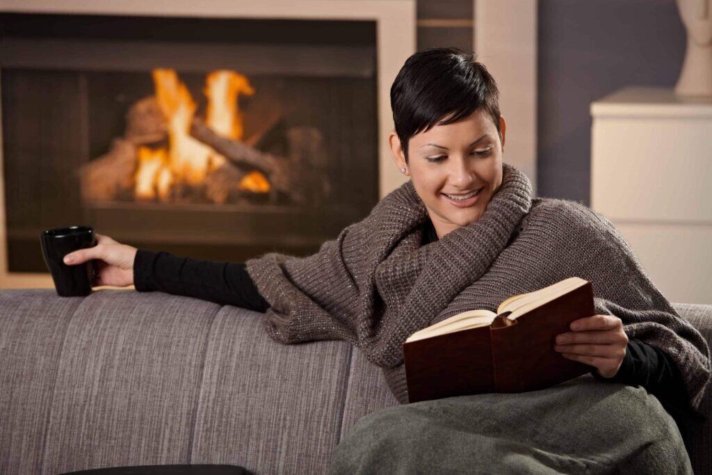 Person sitting on a couch by a fireplace, wrapped in a blanket, holding a mug, and reading a book.