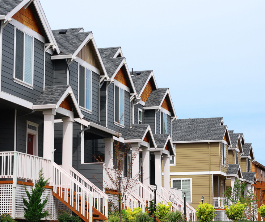 Row of modern townhouses with gray and beige siding, white trim, and peaked roofs. Each has a small front yard and porch, with a clear sky in the background.