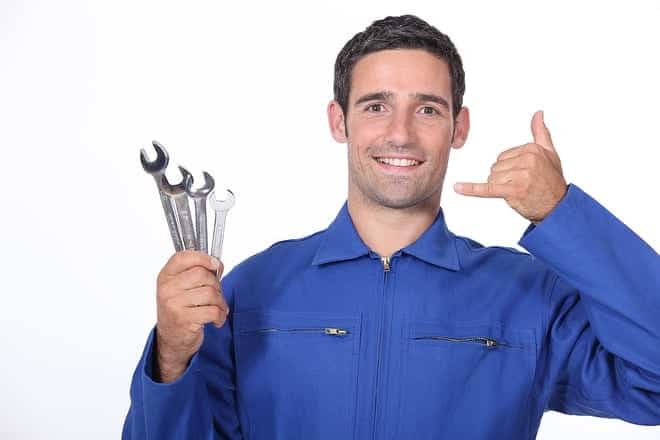 A person in a blue jumpsuit holds three wrenches in one hand and makes a phone gesture with the other hand against a plain white background.