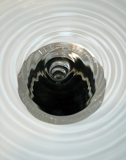 Close-up of a sink drain with water swirling, creating a vortex pattern. The drain is surrounded by reflective metal.