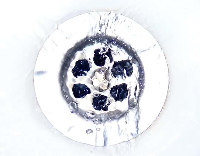 A circular metal drain with small dark debris and flowing water in a white sink.