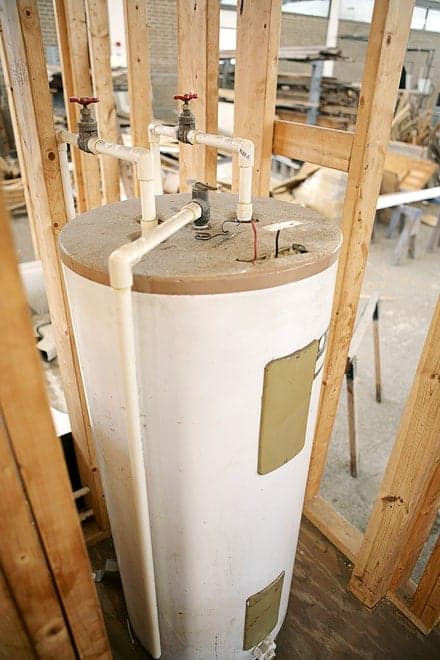 A tall, cylindrical water heater with multiple white pipes and red valves is situated within a wooden frame structure.