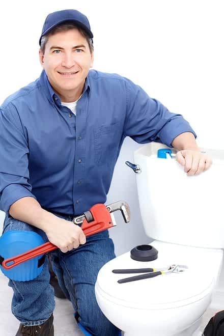 A person kneels beside a toilet, holding a pipe wrench. Other tools like pliers and tape are nearby. The person wears a blue shirt, jeans, cap, and knee pads, smiling at the camera.