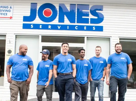 Five men in blue shirts stand smiling outside a building with a sign that reads "JONES SERVICES," showcasing their pride in offering expert general heating repair.