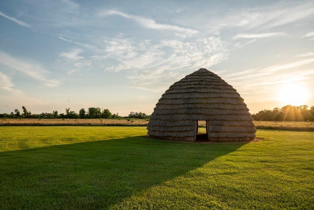 A Caddo Mound sitting atop grass with a bright blue sky and the sun in the background.