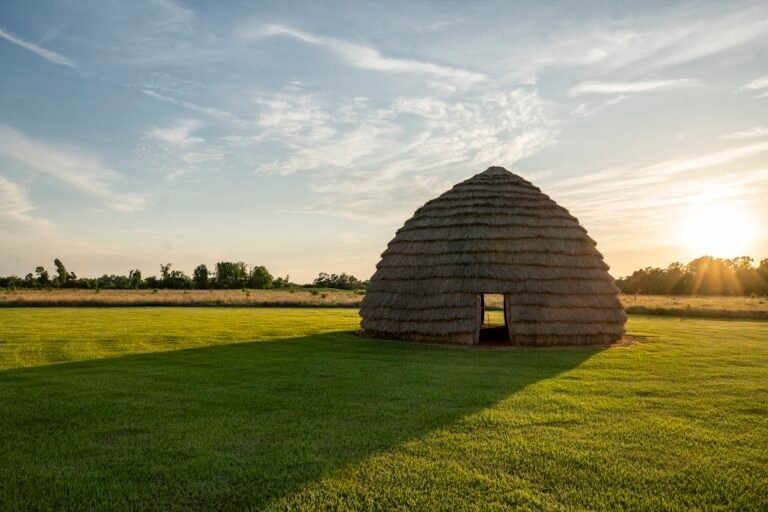 A Caddo Mound sitting atop grass with a bright blue sky and the sun in the background.