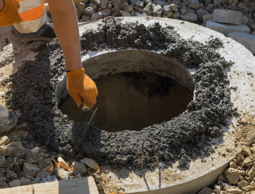 A construction worker wearing orange gloves smooths wet concrete around the circular opening of a manhole—possibly for septic tank maintenance—surrounded by rocks and rubble at a construction site.
