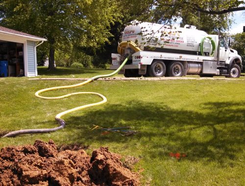 A septic tank truck with hoses attached, used for septic service and grease trap pumping, is parked on a sunny lawn near a house, with dug-up soil and exposed pipes in the foreground among green grass and trees.