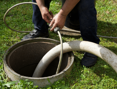 A person is holding hoses near the opening of an outdoor septic tank, preparing for septic system service or cleaning. The surrounding area is grassy.