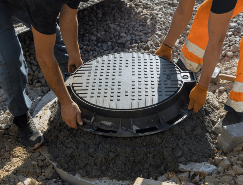 Two workers in orange safety clothing are placing a round manhole cover onto a concrete opening surrounded by gravel at a construction site, performing septic system service to ensure proper maintenance and functionality.