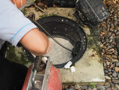 A person uses a plumbing snake to clear a clogged outdoor drain near a septic system, surrounded by stones and debris, wearing a short-sleeved shirt and face mask—important for proper septic tank maintenance.