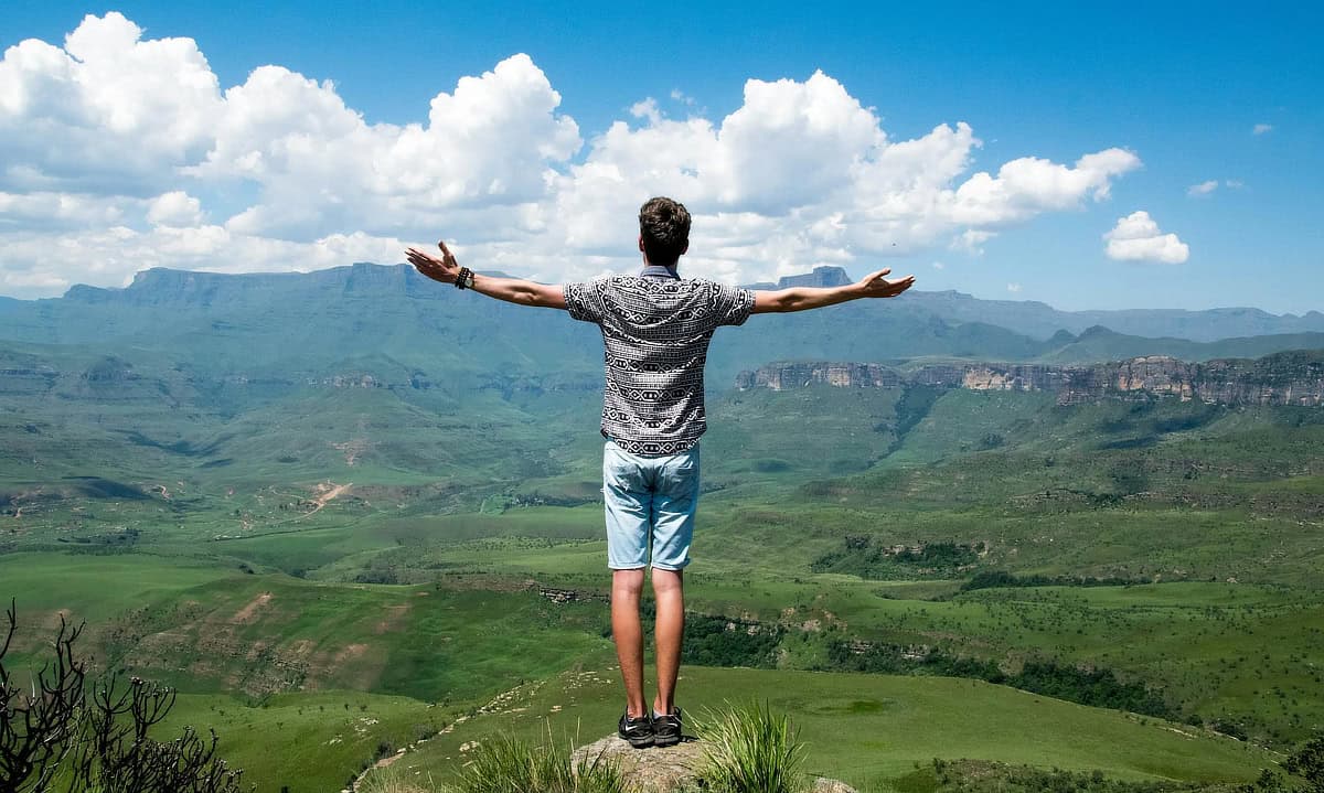Man wearing grey shirt and blue shorts standing on elevated surface with his arms raised in front of a distant mountain view.