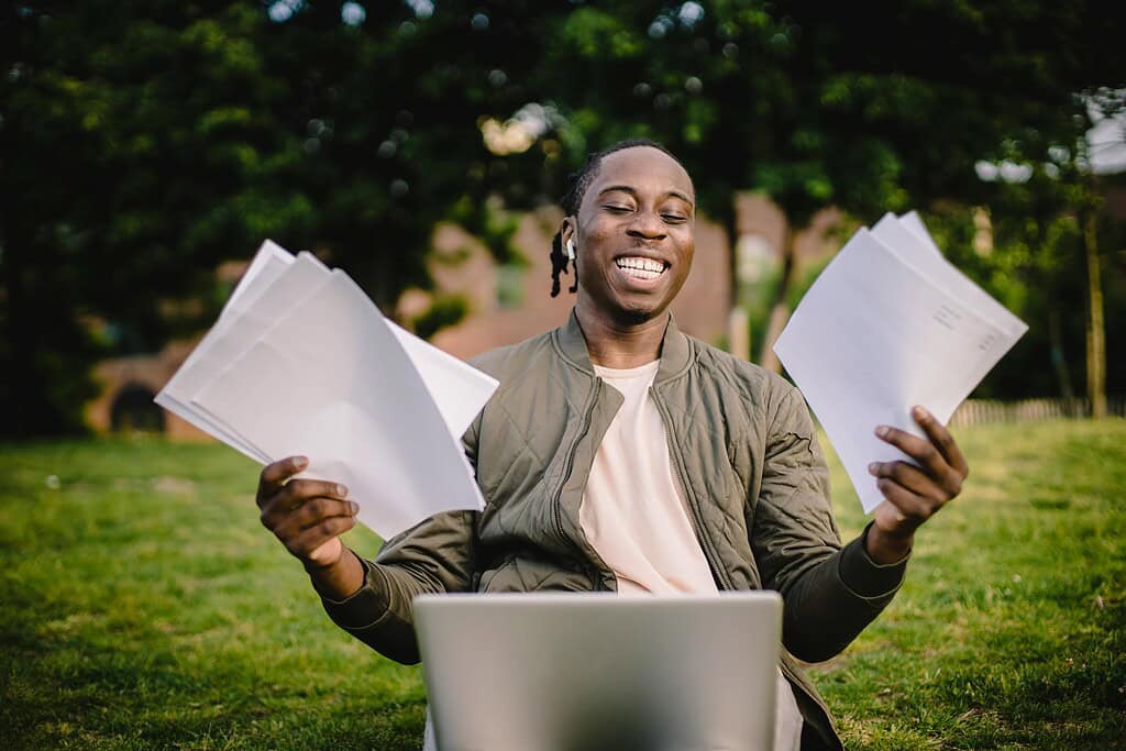 A man celebrating his first freelance writing assignment. He is sitting at a laptop and holding paper in both hands.