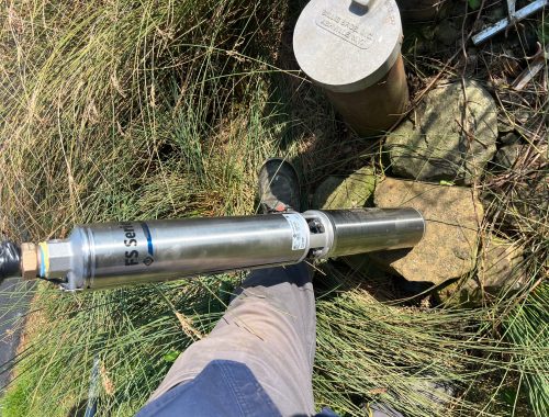 A worker from McCandless Well Drilling and Services Inc. checks a water pump at a well site surrounded by grass and rocks.