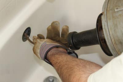 A person wearing gloves uses a plumbing snake to clear a bathtub drain.