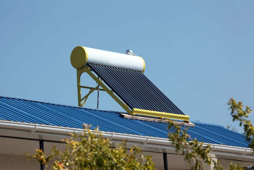 A solar water heater with black tubes and a white tank is mounted on a slanted blue metal roof under a clear sky. Trees partially frame the foreground.