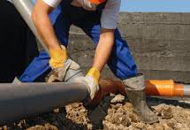 Worker in gloves and safety gear connects large pipes at a construction site, surrounded by gravel and soil.