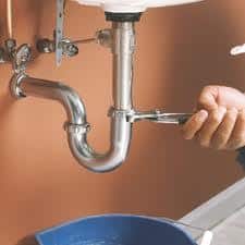 A hand uses a wrench to tighten a pipe under a sink, with a blue container placed below.