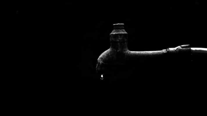 A single drop of water hangs from the spout of an old, rusted outdoor faucet against a dark background.