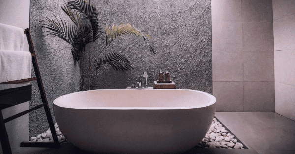 Minimalist bathroom with a white oval bathtub on pebbles, a textured gray wall, a wooden towel rack with white towels, and a potted plant in the corner.