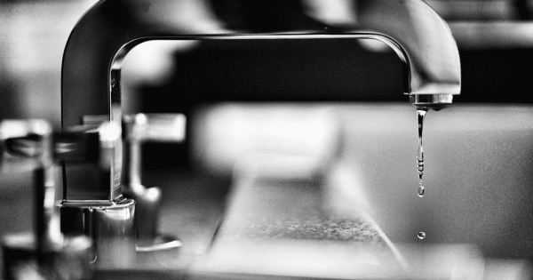 Close-up of a modern faucet with water droplets falling from the spout. The image is in black and white, emphasizing the sleek metal design against a blurred background.
