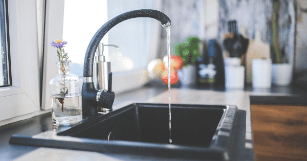 Water flows from a black kitchen faucet into a sink, with a small flower vase and various items on the countertop.