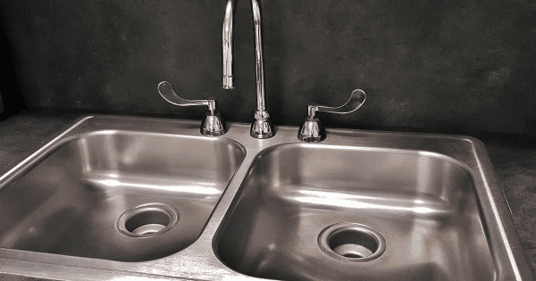 A stainless steel double sink with a chrome faucet and dual handles against a dark backsplash.
