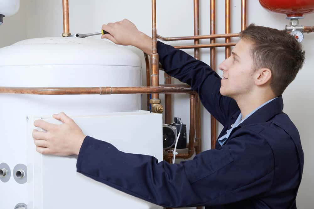 A person in a blue uniform is inspecting a boiler system with copper pipes, adjusting a component with a screwdriver.