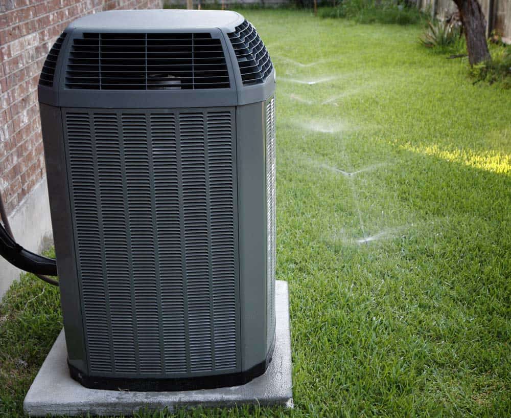 Air conditioning unit on a concrete slab next to a house. It is surrounded by a grassy lawn with sprinklers in operation.