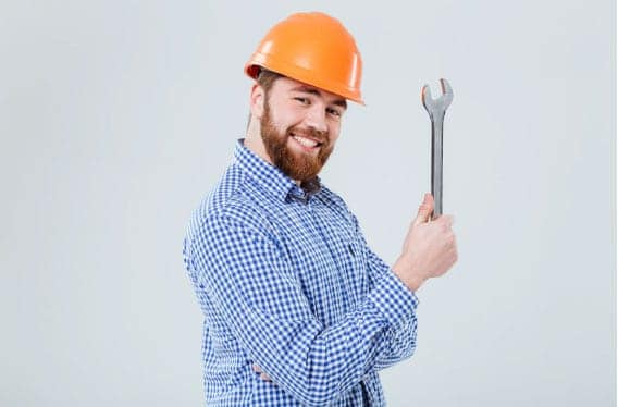 A person wearing an orange hard hat and checkered shirt holds a large wrench, smiling at the camera.