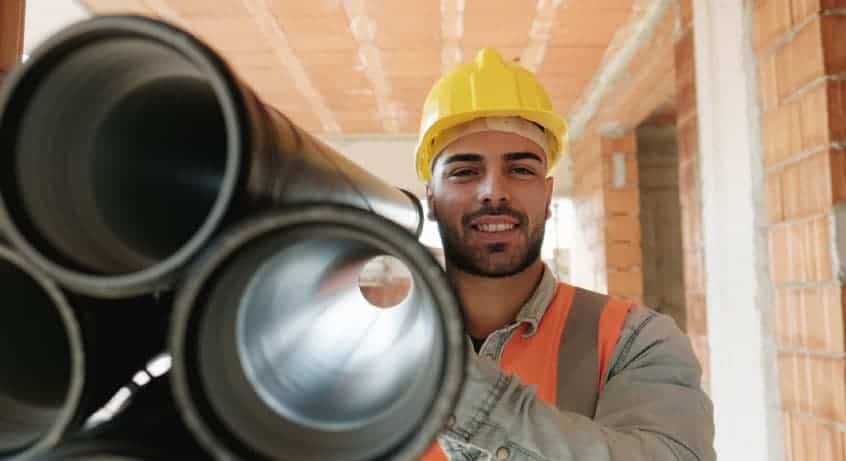 Construction worker in a yellow hard hat and safety vest holds large black pipes, standing in a partially built structure with exposed brick walls.