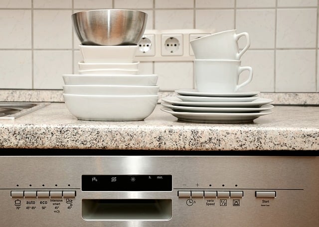 Stacked white bowls, cups, and plates sit on a granite countertop above a stainless steel dishwasher with visible control buttons.