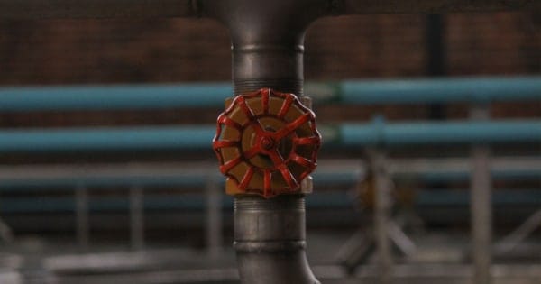Close-up of a metal pipe with a red valve wheel in an industrial setting, showing blue pipes in the blurred background.