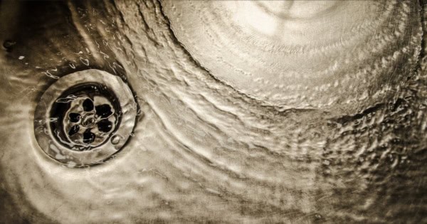 Water spiraling down a metal drain, creating ripples in a textured sink surface, captured in sepia tone.