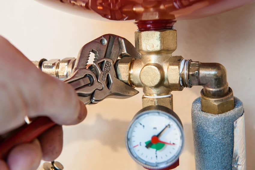 A hand uses an adjustable wrench on a brass plumbing valve. A pressure gauge with a red needle is attached nearby.