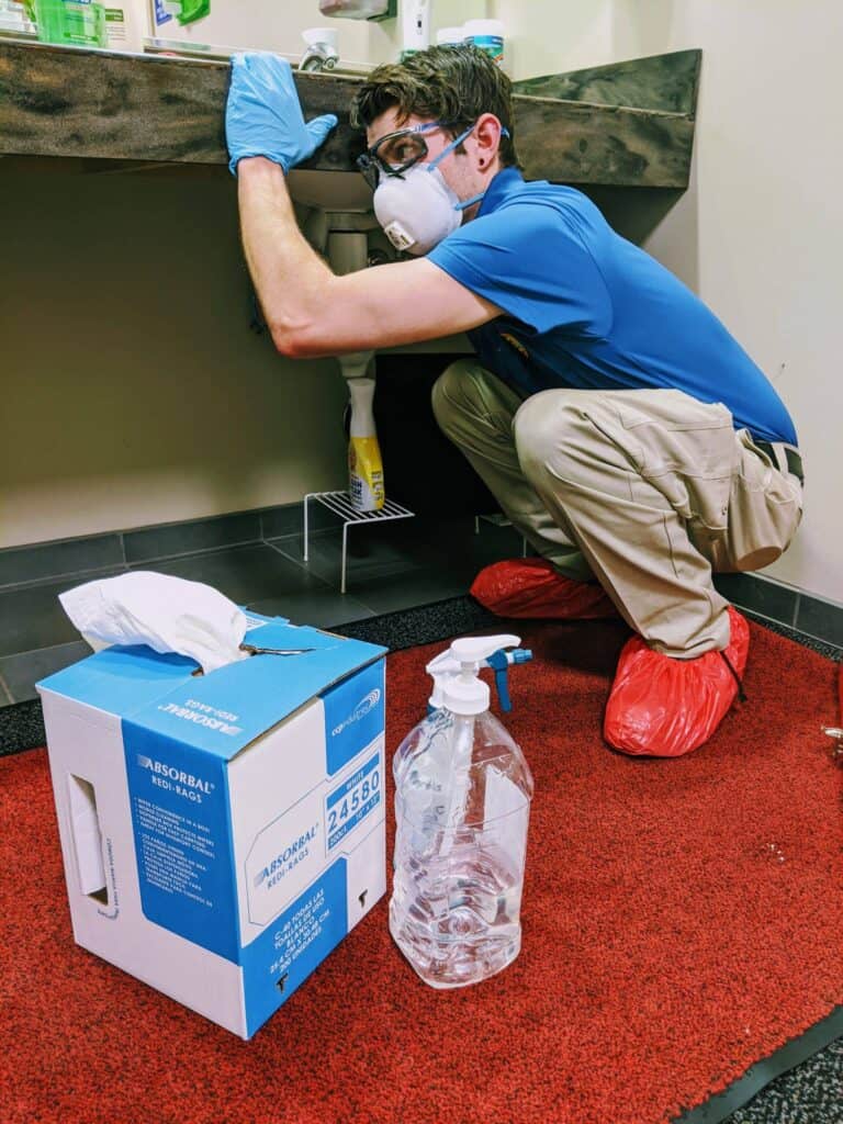 Person wearing protective gear works under a sink. Box of absorbent material and cleaning spray bottle are on a red mat in the foreground.