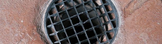 Close-up of a round metal drain cover on a brown surface, featuring a grid pattern allowing water to pass through.