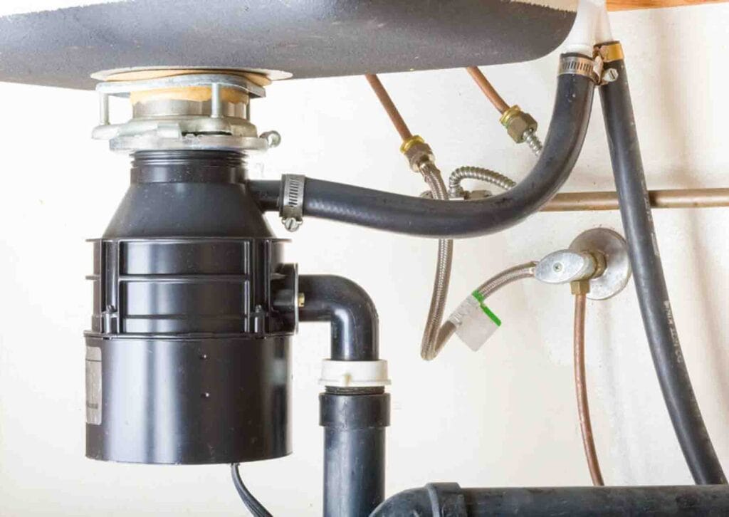 Under-sink view of a garbage disposal unit connected to pipes and hoses, with visible plumbing fixtures and connections.