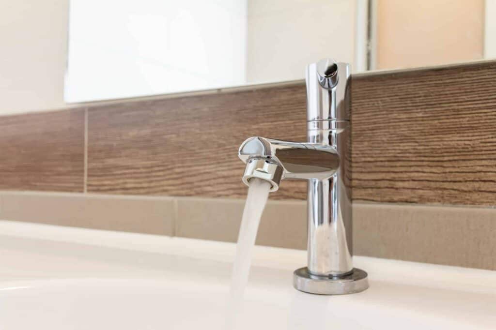 A chrome faucet releases water into a white basin, with a wooden-textured backsplash visible in the background.