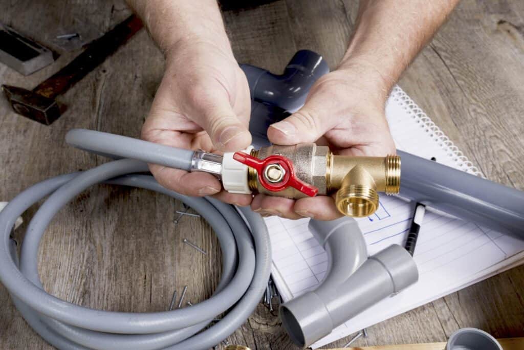 Hands are assembling plumbing components on a wooden table, including pipes, a valve, and a flexible hose. A notebook and pen are in the background.