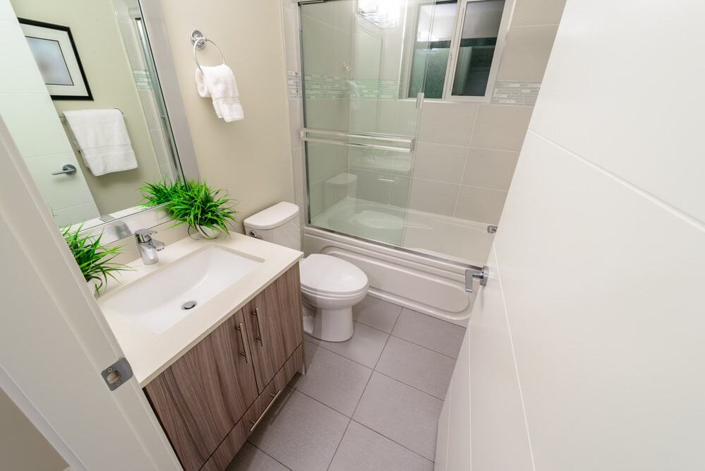 Modern bathroom with a sink, toilet, and bathtub. Beige tiles, glass shower doors, and wooden vanity with a green plant on top. Towels hang near the mirror.