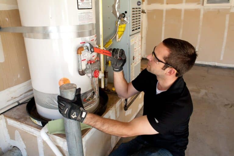 A man wearing gloves uses a wrench to adjust a valve on a water heater in a garage.